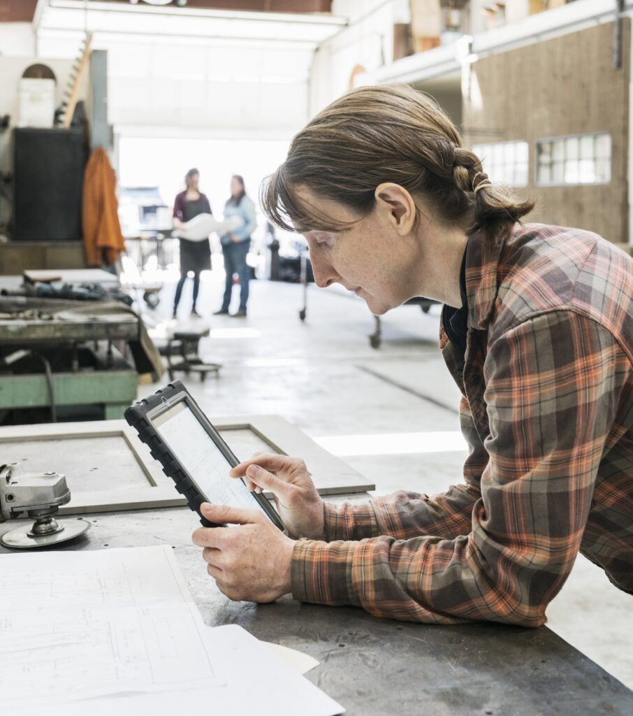 Woman standing at workbench in metal workshop, holding digital tablet.